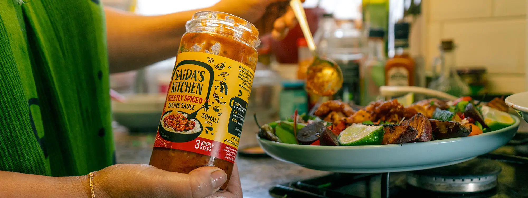 Saida holding a jar of Sweet & Tangy Xawaash (Hawaji) sauce, Somali style, beside a plate of roast vegetables